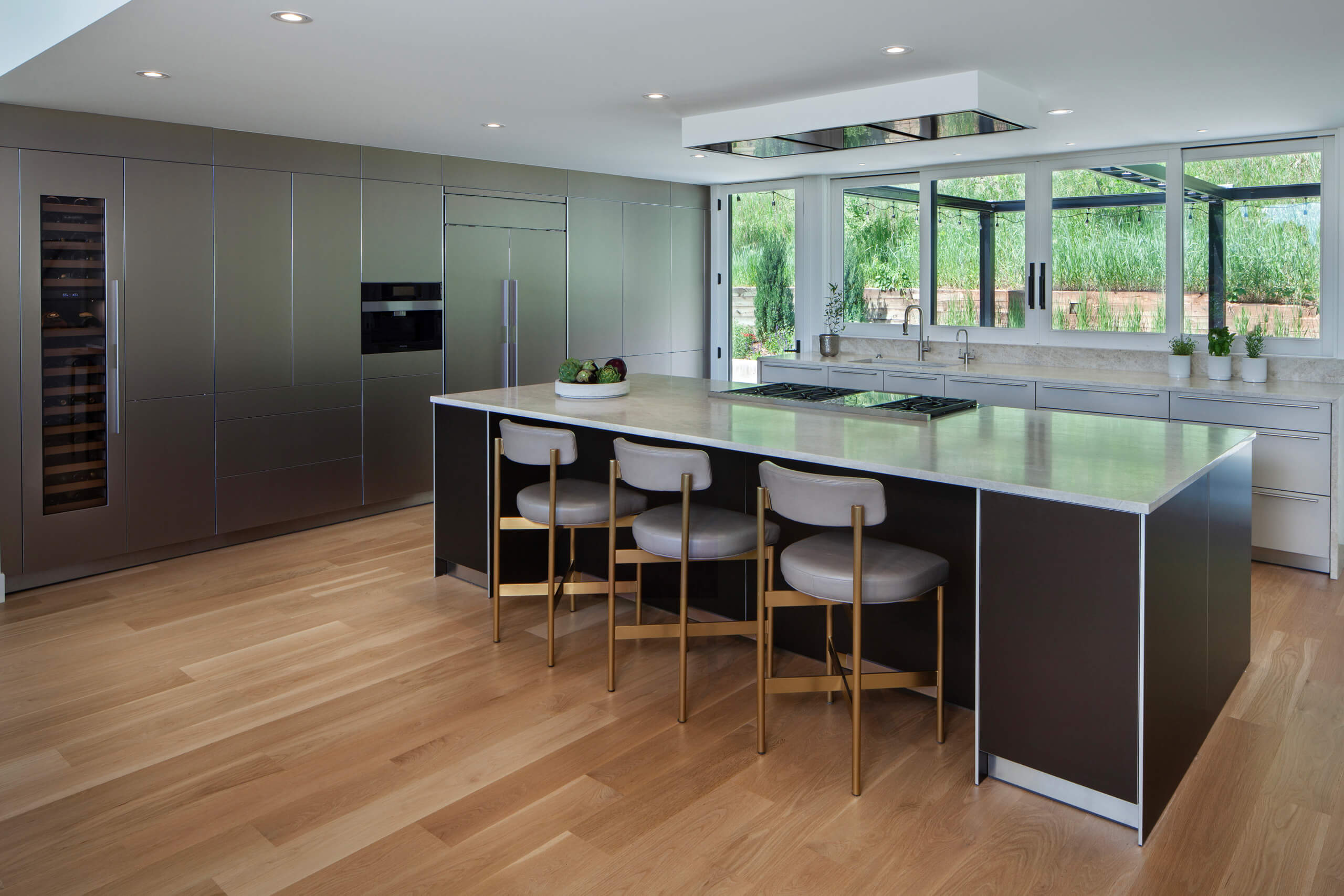 Timeless luxury kitchen with dark cabinetry, wine fridge, kitchen island with gold bar stools, and garden views by Kitchen Distributors