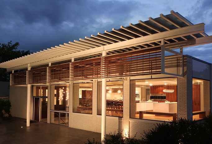 modern-home-wood-slat-pergola-dusk-exterior Bulthaup kitchen showroom storefront at dusk, with large glass windows showing a wood and stainless steel kitchen display, brick base, and metal roof overhang.