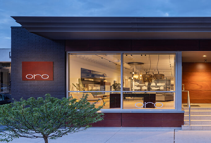 oro-showroom-storefront-dusk-exterior Oro furniture showroom storefront at dusk, featuring dark brick facade, illuminated glass windows revealing modern furniture and pendant lighting, and an angular metal roof canopy.